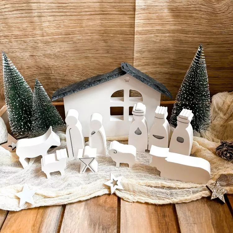 A imagem retrata um presépio minimalista com figuras brancas de madeira, incluindo a Sagrada Família, Reis Magos e animais. Ao fundo, uma cabana estilizada e pequenas árvores de Natal completam a composição. A cena está disposta sobre uma superfície de madeira, decorada com estrelas e tecido claro.


