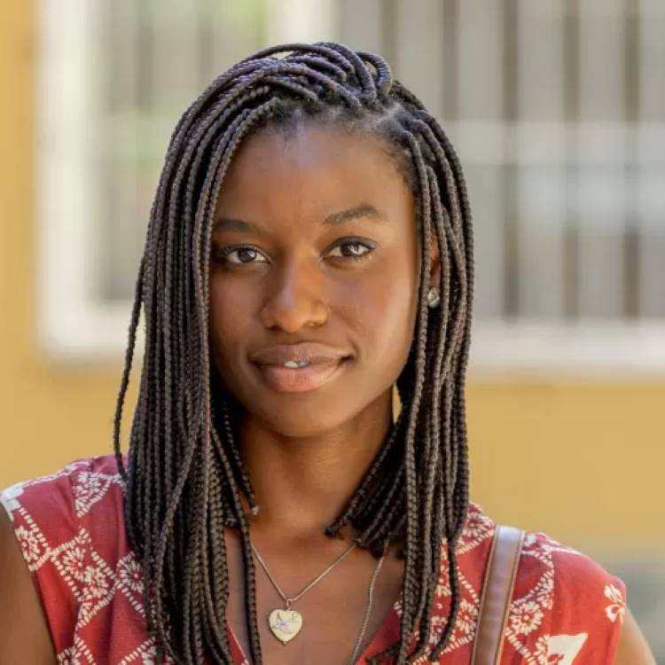 Uma mulher com box braids escuras até os ombros e um brinco de estrela prateado na orelha direita sorri suavemente para a câmera. Ela usa um colar com um pingente de coração dourado sobre uma blusa estampada em tons de vermelho e branco. O fundo é um edifício em tom claro.







