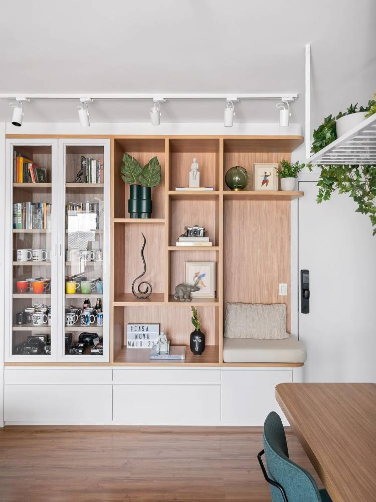 Estante com partes vazadas e alguns nichos fechados, além de um espaço para sentar. Nela, há livros, vasos de planta, quadros e outros itens decorativos. 