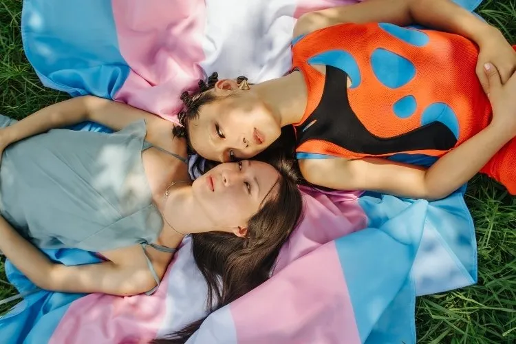 Foto de duas pessoas trans deitadas sobre bandeira azul, rosa e branca