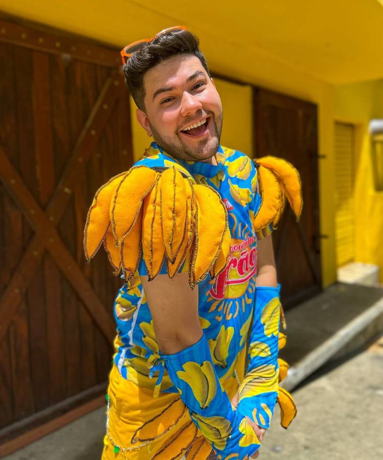 A imagem mostra um homem sorridente vestindo um abadá customizado azul com estampas de bananas amarelas. O design inclui ombreiras volumosas e luvas combinando. Ele está em um ambiente ensolarado, com paredes amarelas e portas de madeira ao fundo.