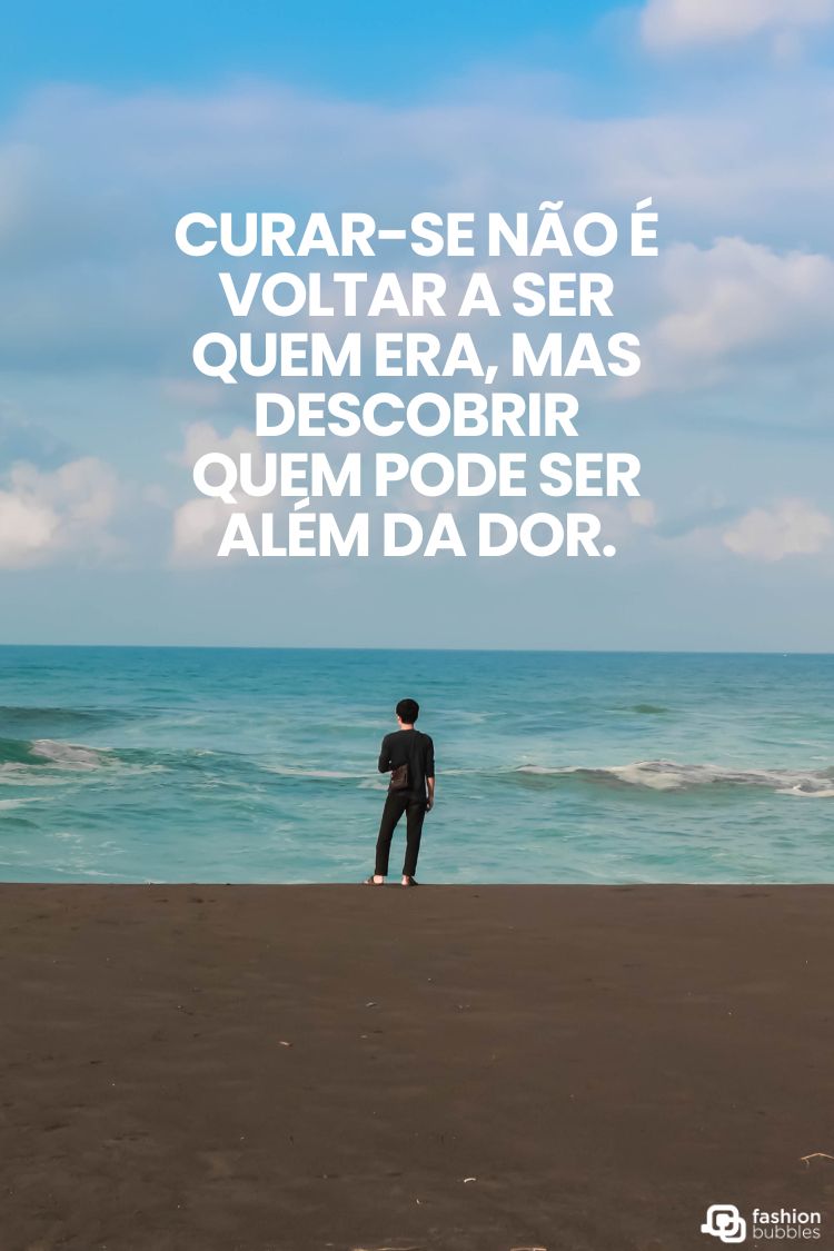 A imagem mostra uma figura solitária de costas, olhando para o vasto oceano sob um céu azul claro, em uma praia escura. A frase "CURAR-SE NÃO É VOLTAR A SER QUEM ERA, MAS DESCOBRIR QUEM PODE SER ALÉM DA DOR" está sobreposta. A foto transmite uma sensação de contemplação e renovação.
