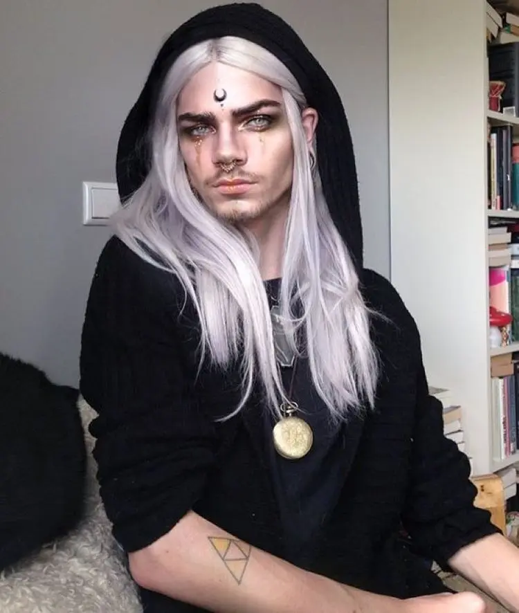 Pessoa de pele clara usando cabelo branco, capuz preto, maquiagem com strass dourado e desenho de lua na testa