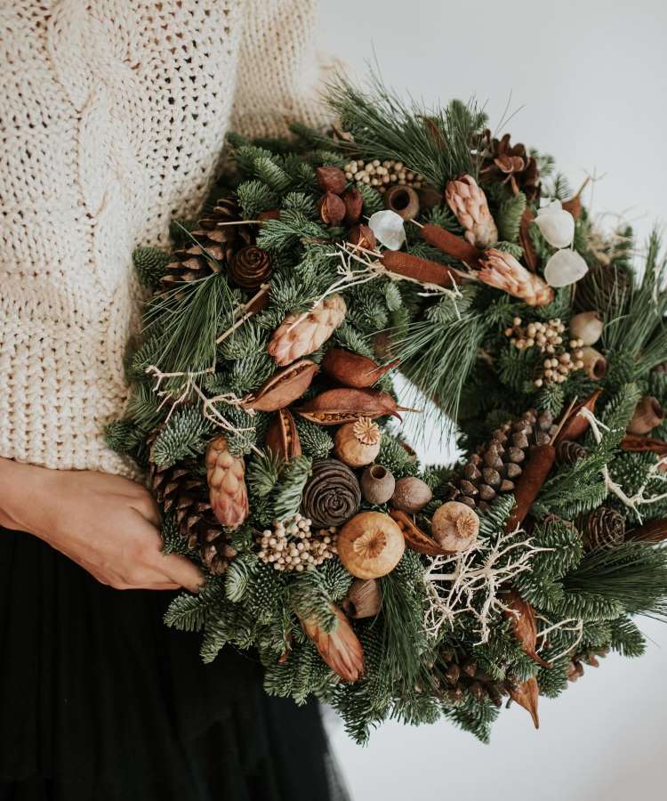 Uma pessoa segurando uma guirlanda de Natal feita com galhos de pinheiro, pinhas, canela e elementos naturais secos. Ela veste um suéter de tricô creme e uma saia preta. A guirlanda tem um visual rústico e elegante, ideal para decoração.


