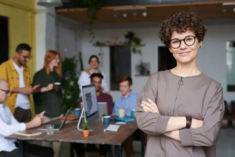 Mulher de jaqueta marrom e óculos, com braços cruzados. Ao fundo, vemos uma equipe de trabalho implementando espiritualidade nas empresas