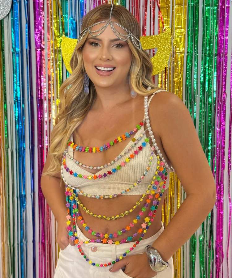 Mulher loira sorridente com cabelo ondulado veste top branco decorado com colares coloridos e calça branca. Usa acessório prateado na cabeça e bracelete. Fundo com cortina brilhante multicolorida e asas amarelas simboliza festa e alegria.
