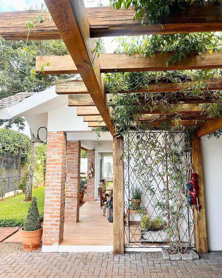 Imagem de fachada de casa simples chique, com colunas em tijolinho, árvores e pergolado com planta trepadeira
