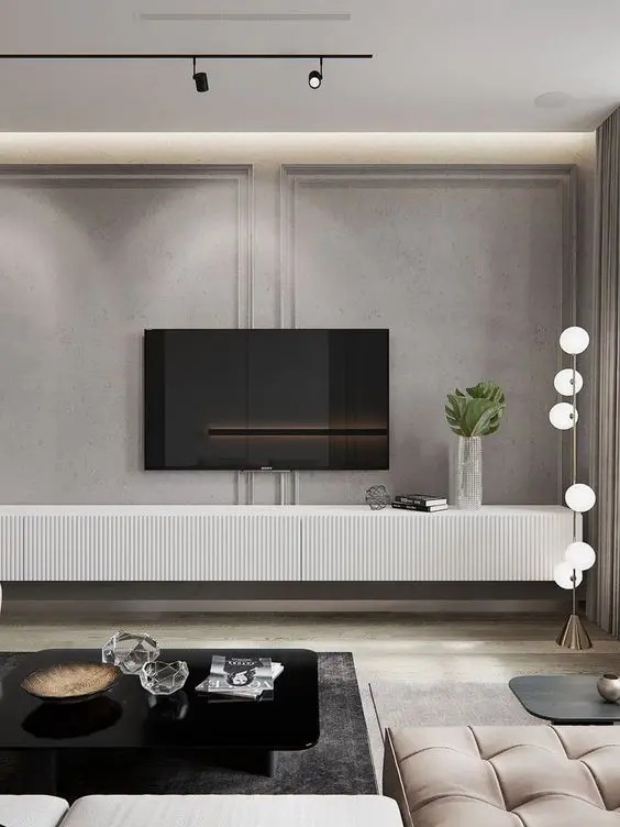 Sala de TV com paredes cinzas com moldura, televisão, aparador branco com livros e vasos, mesa de centro preta com decoração, luminária de pé e sofá bege