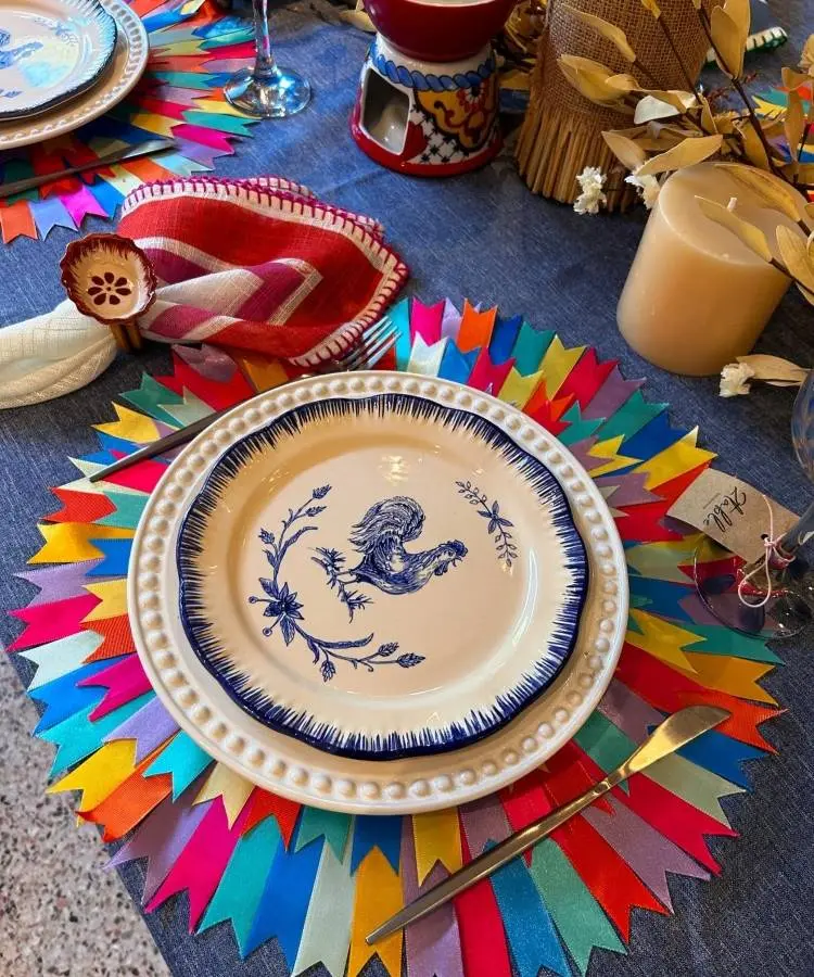 Uma mesa posta com um prato branco decorado com um galo azul e borda com bolinhas. O prato está sobre um sousplat feito de fitas coloridas, e há um guardanapo vermelho e talheres ao lado.