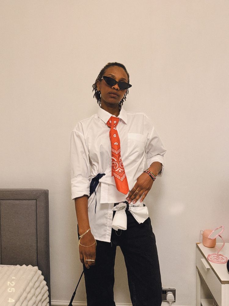Mulher de pele negra com camisa desconstruída branca, bandana laranja e calça preta