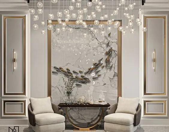 Sala de estar com lustre com diversas lâmpadas, parede branca com boiserie branco e interno dourado, quadro de mármore e duas poltronas beges e cinzas