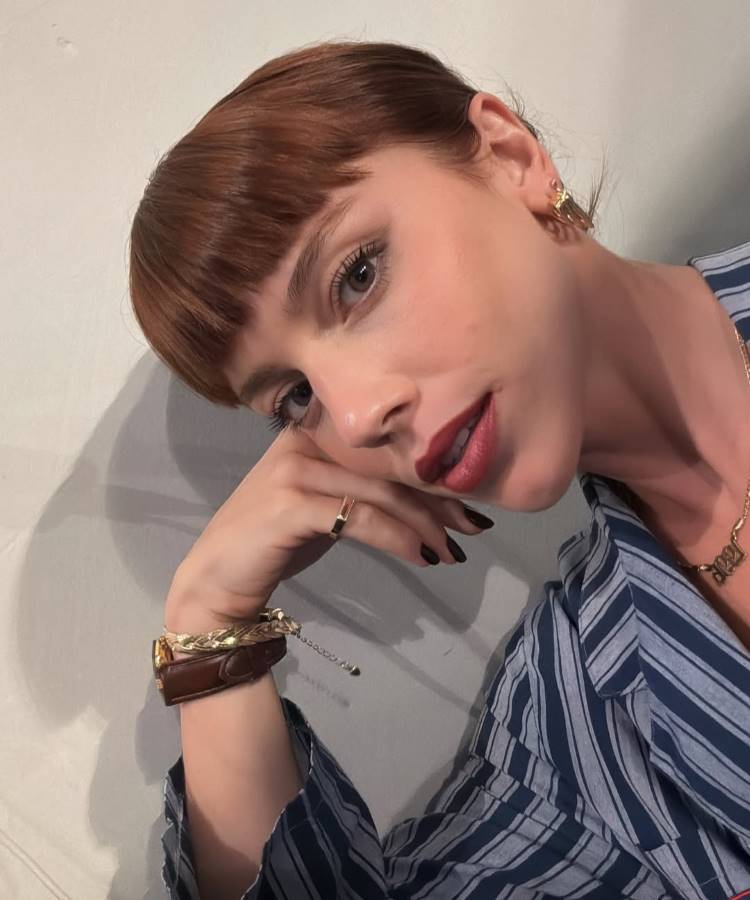Solange (Alice Wegmann) de cabelos ruivos e franja curta, com batom vermelho escuro, apoia a cabeça na mão. Ela usa uma camisa listrada, brincos, um colar com o ano "1996", e joias de Vale Tudo como um anel e pulseira grossa, com unhas pretas.