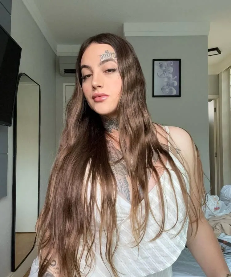 Mulher com longos cabelos castanhos soltos posa em quarto iluminado. Usa vestido branco decotado e mostra tatuagens no peito e pescoço. Na testa, tatuagem em destaque. Expressão confiante, ambiente ao fundo exibe cama desarrumada, parede cinza e quadro decorativo.