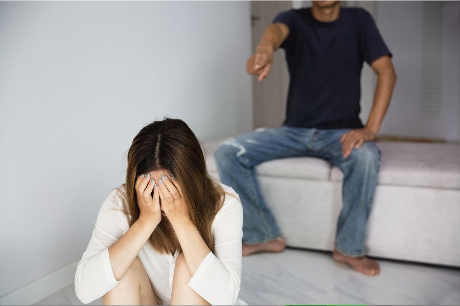 foto de mulher sentada no chão com as mãos no rosto enquanto um homem briga com ela, simbolizando mulheres que voltam para agressores