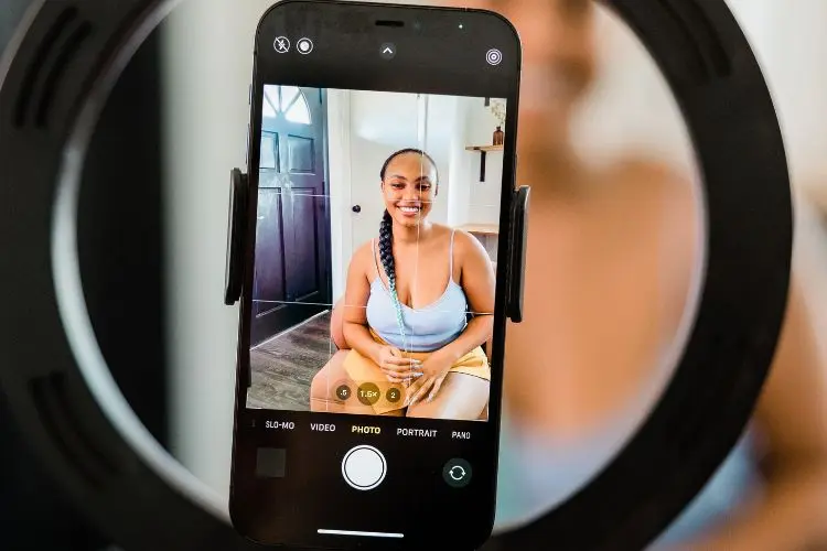 A imagem mostra um smartphone em um suporte circular de ring light. Na tela, uma mulher sorridente, de cabelo trançado, vestindo uma blusa azul, está posando. O ambiente é iluminado, com uma porta ao fundo e detalhes decorativos.