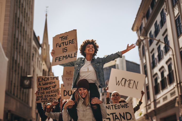 Mulher carregando outra mulher no ombro que segura uma placa escrito o futuro é feminino em inglês. atrás dela, há outras mulheres segurando outras placas em inglês. ao fundo prédios. elas estão andando em uma rua
