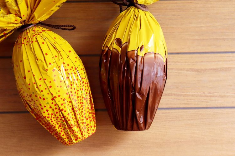 Dois ovos de Páscoa embalados em papel amarelo estão sobre uma superfície de madeira. Um deles possui detalhes em estrelas vermelhas, enquanto o outro tem uma metade com acabamento em chocolate visível na embalagem.
