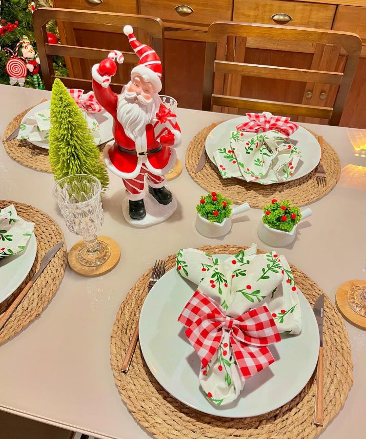 Mesa decorada para o Natal com sousplats de palha, pratos brancos, guardanapos estampados, laços xadrez, taças de vidro e enfeites verdes. Destaque para figura de Papai Noel central.