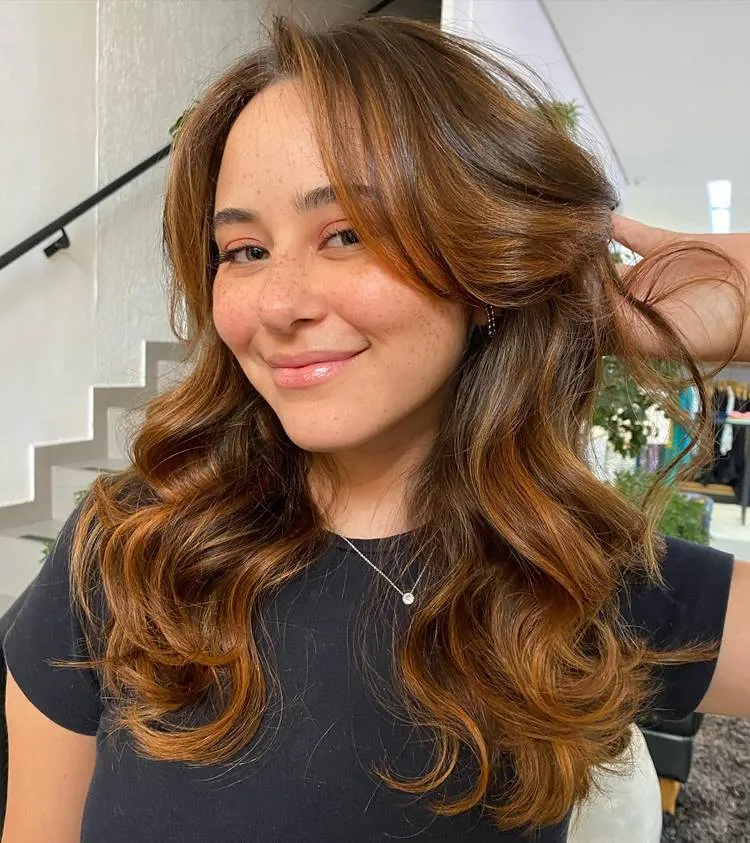 Mulher de rosto redondo e cabelo iluminado, com butterfly bangs. Ela usa camiseta preta