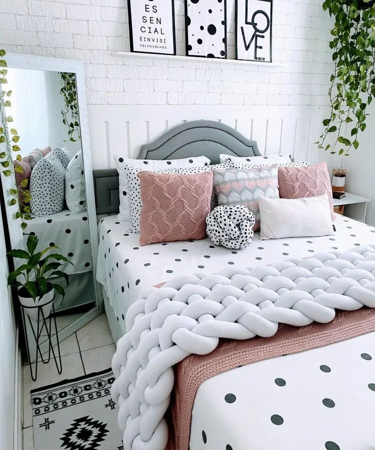 A imagem mostra uma decoração fácil para quarto, com uma cama de casal centralizada, adornada com várias almofadas de diferentes padrões e cores suaves como cinza, rosa e branco. Acima da cama, há quadros decorativos. Um espelho grande e plantas verdes completam o ambiente acolhedor e estiloso.


