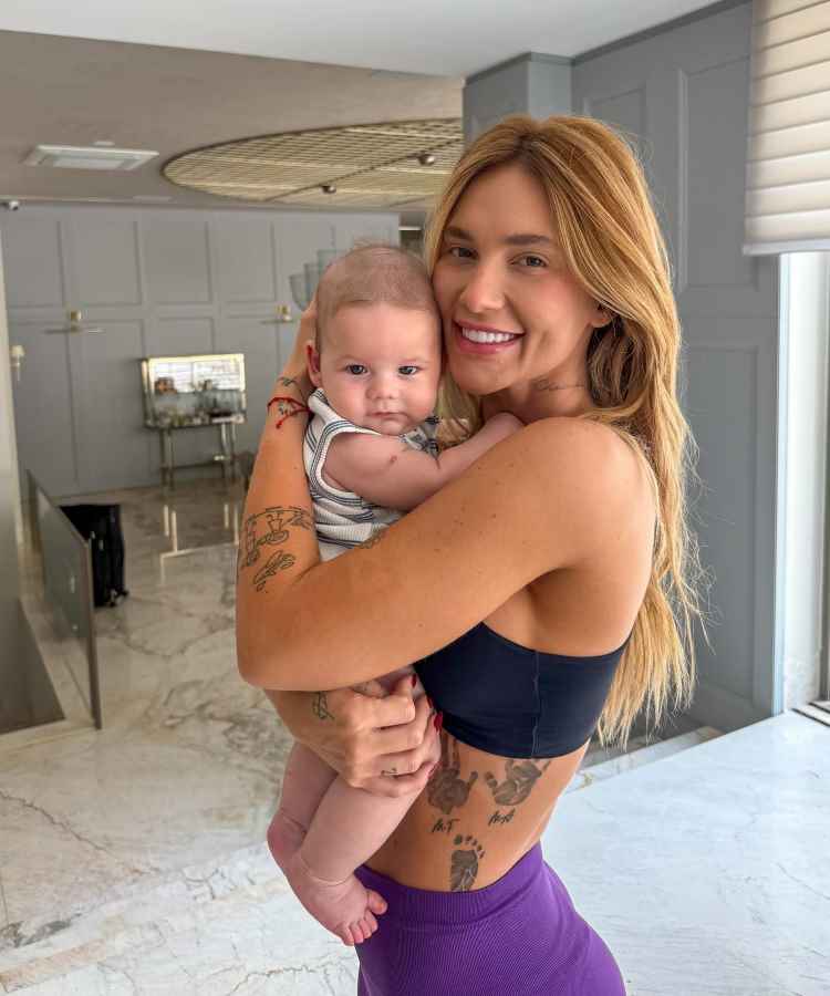 A imagem mostra uma mulher sorrindo enquanto segura um bebê no colo, em um ambiente interno bem iluminado. Ela exibe tatuagens nos braços e abdômen, e usa um top preto e calça roxa. O bebê aparenta tranquilidade.