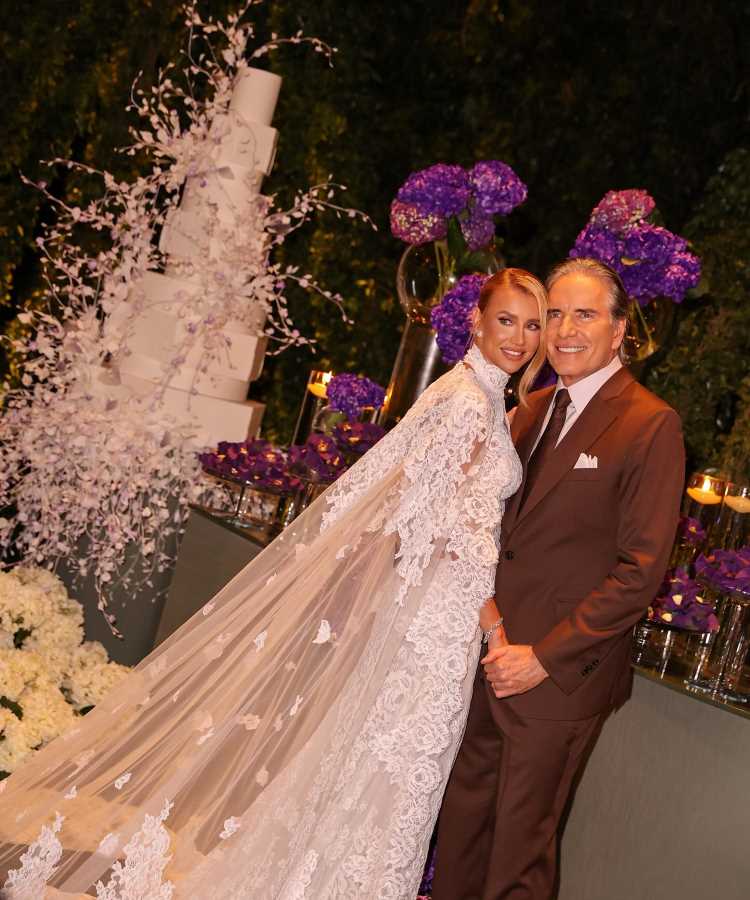 Casal Ana Paula Siebert e Roberto Justus elegante posa em frente à decoração luxuosa com flores roxas e bolo branco em seis andares. Noiva usa vestido rendado com capa longa, enquanto noivo veste terno marrom, destacando o glamour da cerimônia noturna.
