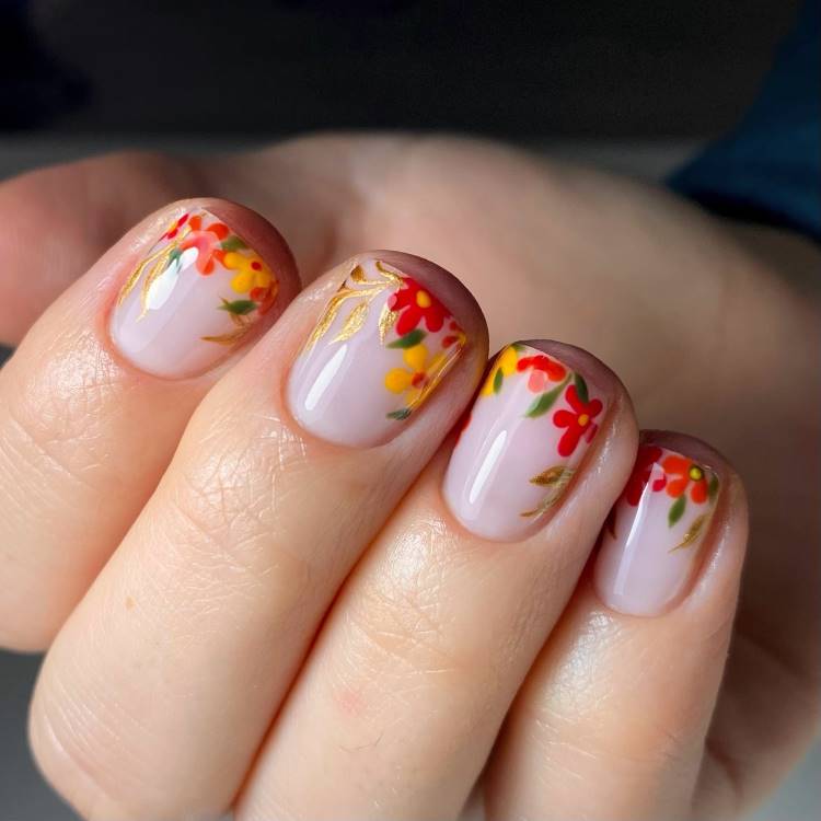 Unhas curtas com esmalte nude decoradas com pequenas flores vermelhas, amarelas e folhas verdes, complementadas por delicados detalhes dourados. O design vibrante e artístico contrasta com o fundo claro, conferindo um toque elegante e primaveril às mãos.





