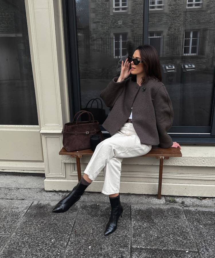 Mulher posa sentada em banco de madeira, usando jaqueta marrom, calça branca e botas pretas de couro. Segura óculos escuros, com bolsa estruturada marrom ao lado. Cenário urbano discreto inspira looks de trabalho casuais e sofisticados para dias frios.





