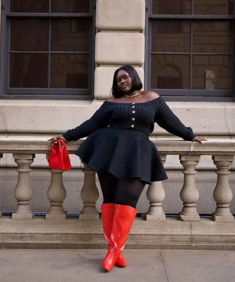 Uma mulher sorridente com cabelo curto e escuro veste uma blusa de tricô preta de ombro a ombro e uma saia preta rodada. Ela usa meia-calça preta e botas vermelhas vibrantes de cano alto, posando em frente a um corrimão de pedra com janelas ao fundo.