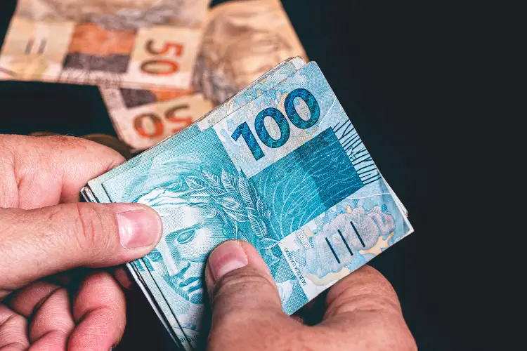 A imagem mostra mãos segurando várias notas de cem reais em primeiro plano. Ao fundo, notas de cinquenta e cem reais estão parcialmente visíveis, contra um fundo preto.


