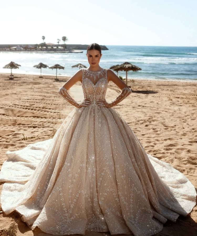 Uma noiva posa na praia, usando um deslumbrante vestido de noiva brilhante, com decote transparente, mangas compridas e saia volumosa. O mar azul, guarda-sóis e palmeiras emolduram o fundo ensolarado.







