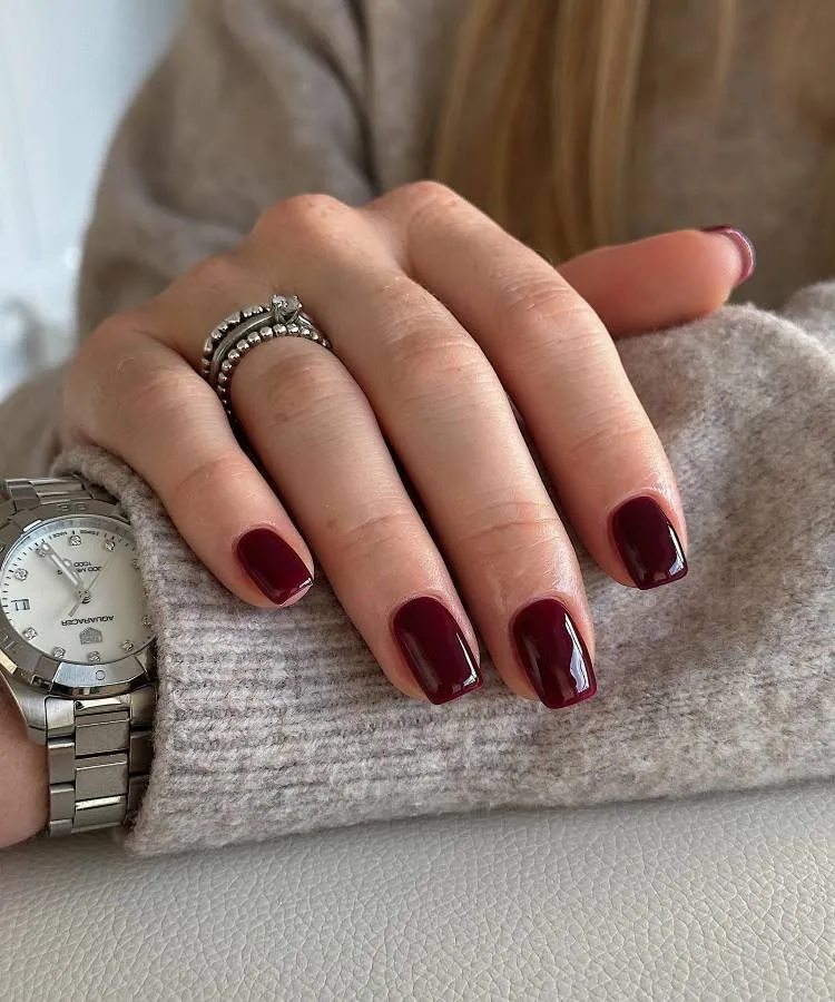 Unhas elegantes no formato quadrado com a ponta arredondada na cor vermelho cereja. A mão com o anel está apoiada no braço da pessoa que usa um casaco de lã e um relógio.
