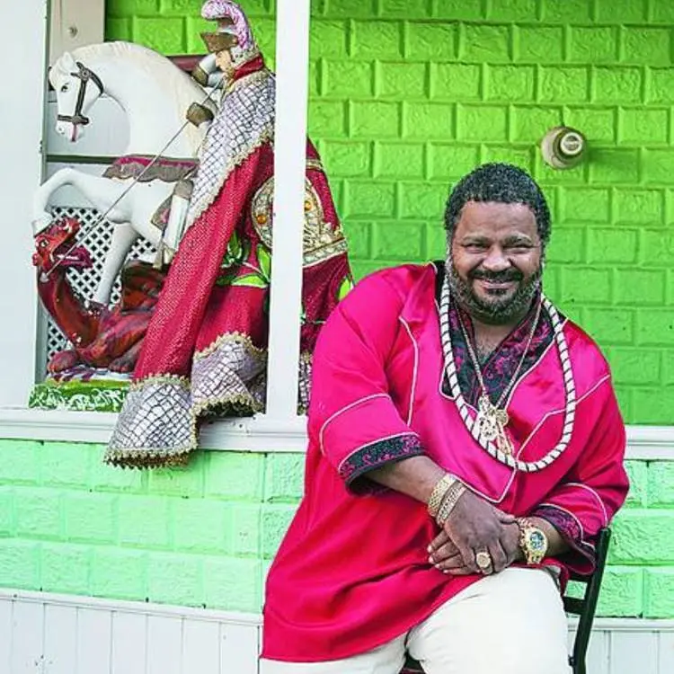 Cantor Arlindo Cruz antes de sofrer o AVC, sentado em cadeira ao lado de imagem de São Jorge