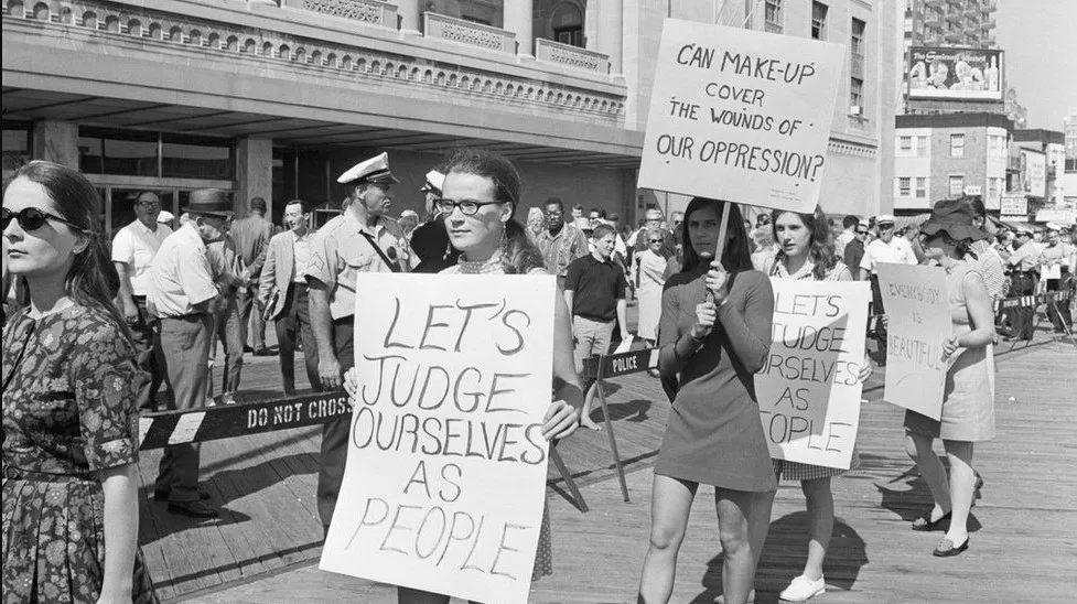 Foto em preto e branco de uma passeata de mulheres em um protesto em 1968 contra o concurso Miss America em Nova Jersey, onde se lê "Vamos nos julgar como pessoas" e "Pode a maquiagem disfarças as feridas da opressão?". Passeata de mulheres em um protesto em 1968 sobre a moda e cidadania contra o concurso Miss America em Nova Jersey, onde se lê "Vamos nos julgar como pessoas" e "Pode a maquiagem disfarças as feridas da opressão?".
