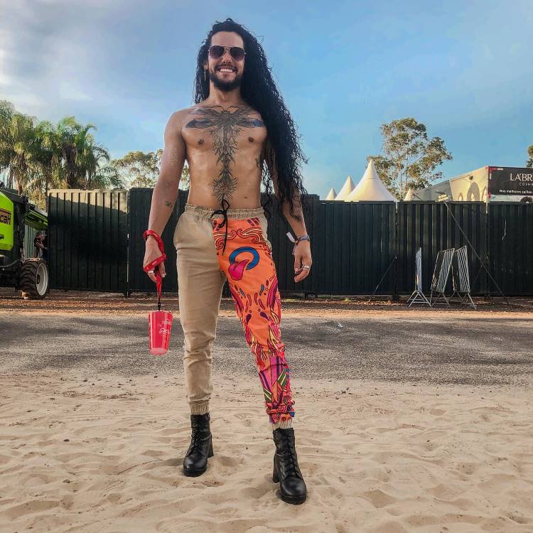 A imagem mostra um homem sorridente, de cabelos longos, óculos escuros e tatuagens no torso. Ele veste calça assimétrica, com uma perna bege e outra colorida, segura um copo vermelho e está em um ambiente externo ensolarado.