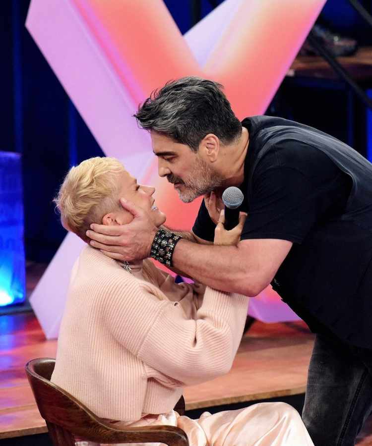 Xuxa sentada em cadeira bem próxima de Junno Andrade, seu esposo, no Altas Horas Especial - Xuxa 60 anos.