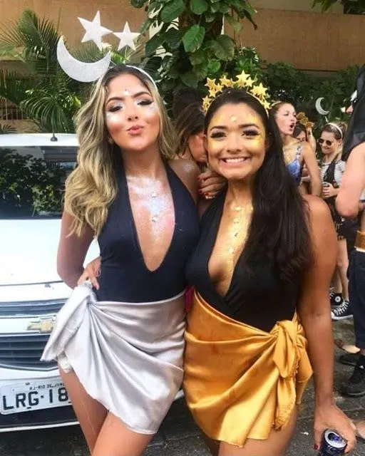 Duas mulheres de body preto, mas uma usa saia prata e tiara de lua e estrelas e outra usa saia dourada com tiara de sol