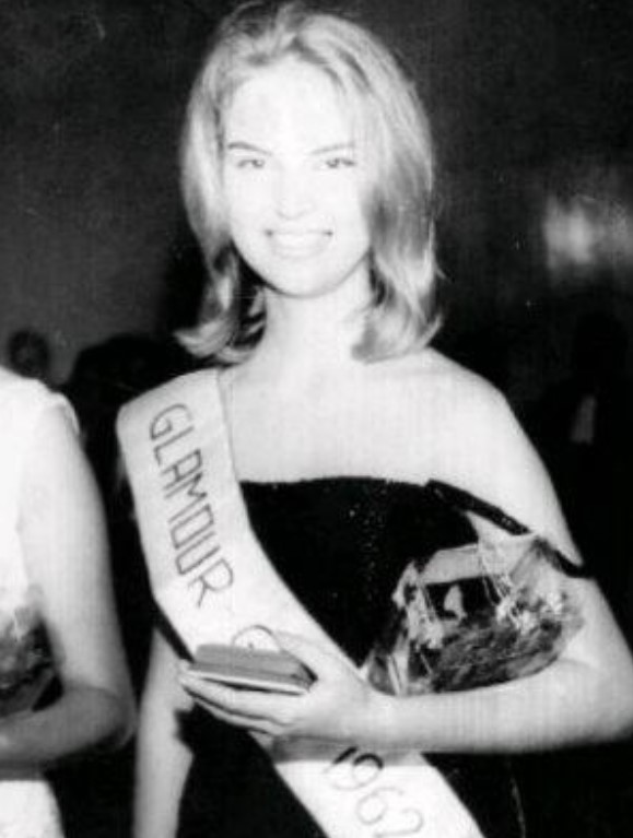 Foto em preto e branco de Elke Maravilha jovem, usando um vestido preto e sorrindo com a faixa do concurso Glamour Girl 1962. Foto em preto e branco de Elke Maravilha jovem, com um vestido preto e sorrindo ao usar uma faixa de Glamour Girl 1962.