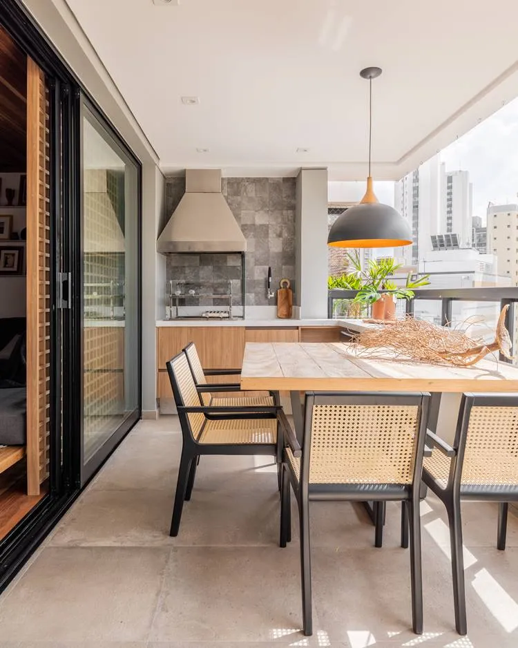 Varanda do apartamento decorado de 187 m². Mesa de 4 cadeiras, com luminária preta e bronze. De decoração no centro, um tronco enraizado seco. Ao fundo, uma churrasqueira, ao lado objetos de cozinha, como faca e tábua