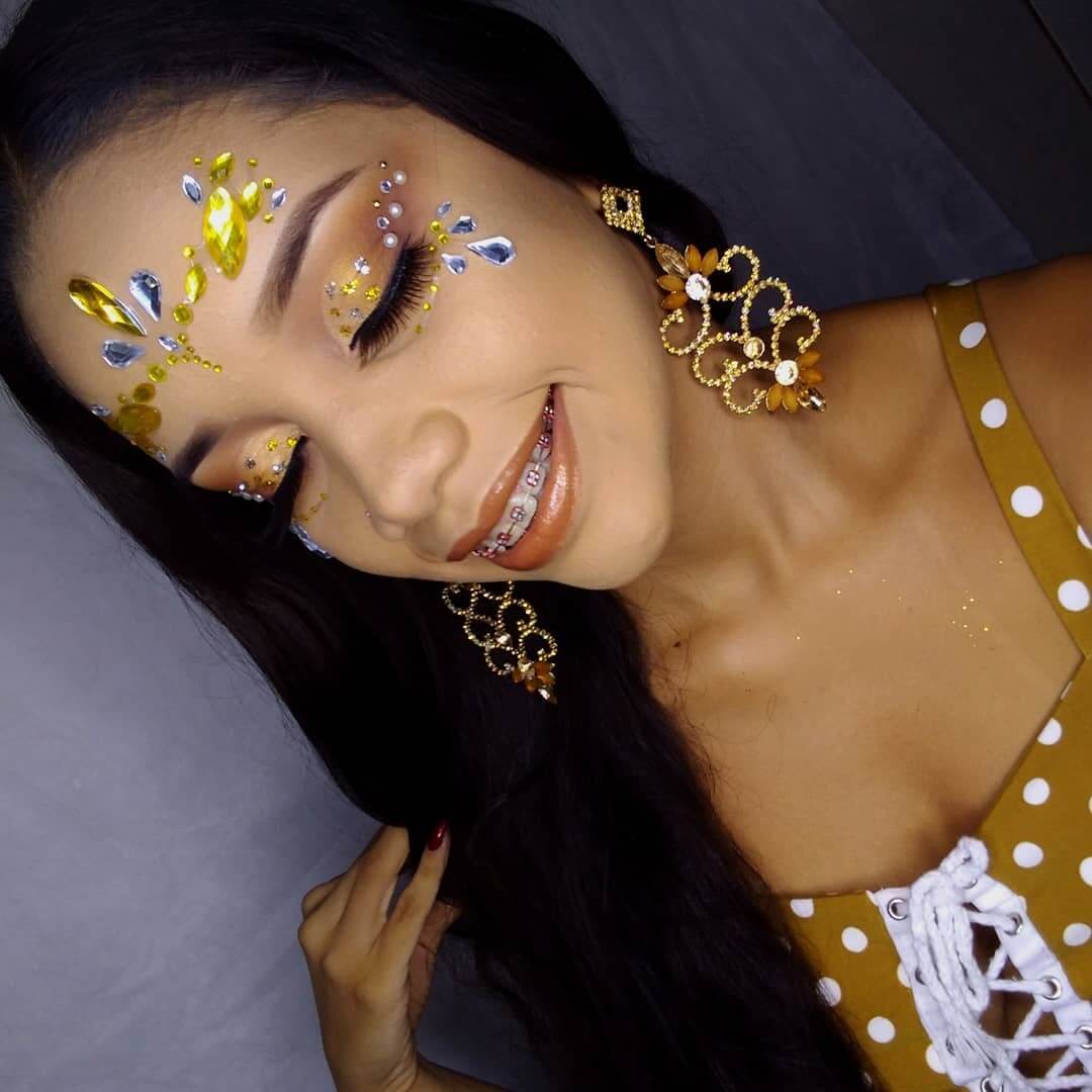 Mulher de pele clara com maquiagem dourada, pérolas, gotas prateadas, folha de ouro e pedrarias em dourado e prateado na testa 