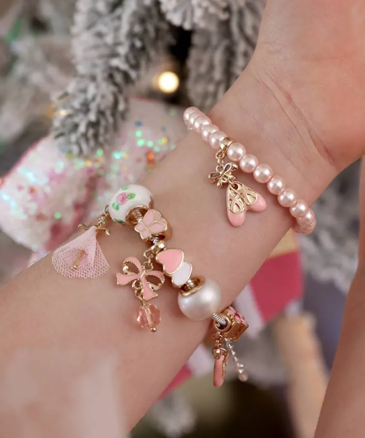 Um pulso infantil usando duas pulseiras: uma com pérolas e um pingente de sapatilha de balé e outra com berloques variados, incluindo um laço rosa, um coração e detalhes de balé.

