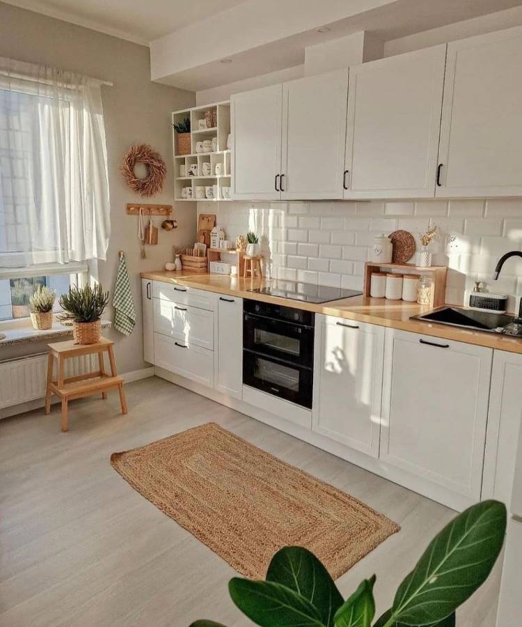 Cozinha com decoração Feng Shui, organizada e iluminada por luz natural. Armários brancos, prateleiras abertas, bancada de madeira clara e tapete de fibra promovem harmonia. Toques naturais com plantas, cortinas leves e elementos terrosos criam energia equilibrada e acolhedora.