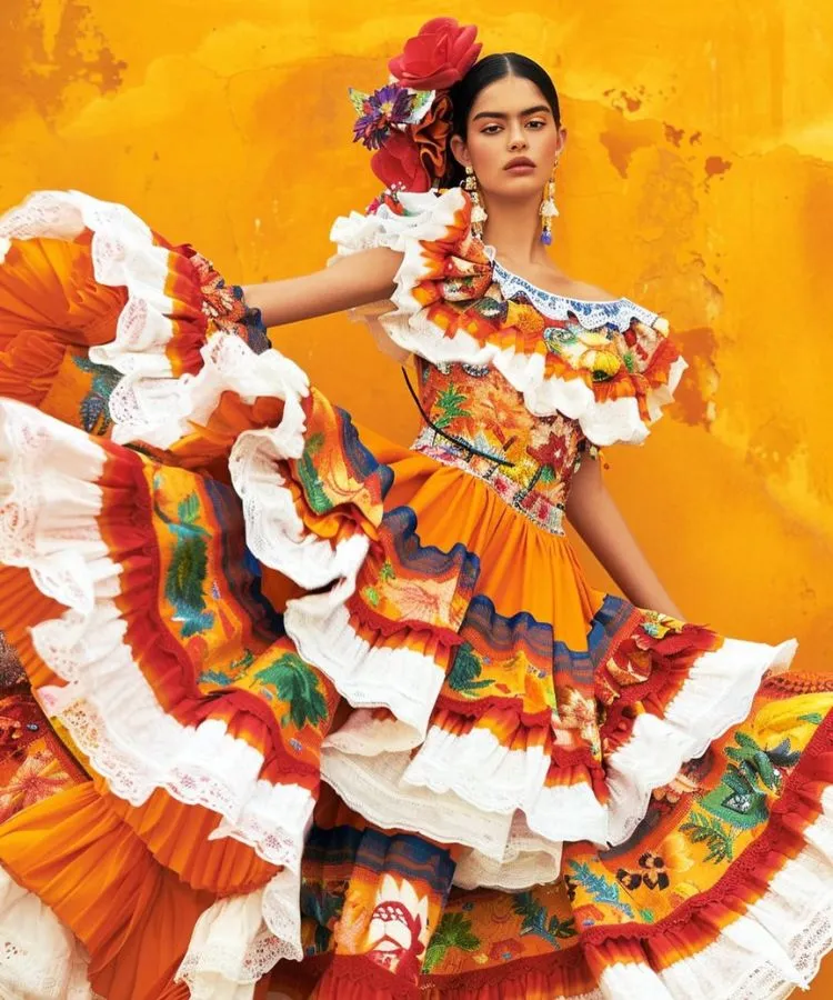 Uma modelo, possivelmente retratando uma dançarina mexicana, posa com um vestido de babados laranja e branco. O vestido tem detalhes florais e babados em camadas. O cabelo da modelo está preso com flores e ela usa brincos pendentes, em frente a uma parede amarela.

