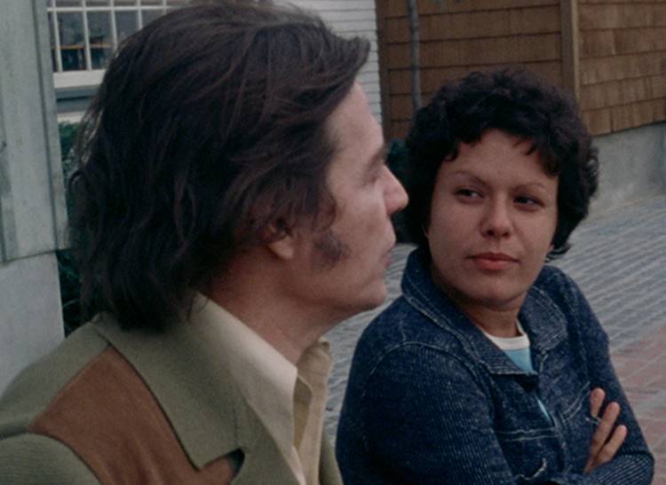 Foto antiga dos músicos Elis Regina e Tom Jobim, eles estão sentados em calçada e se olhando