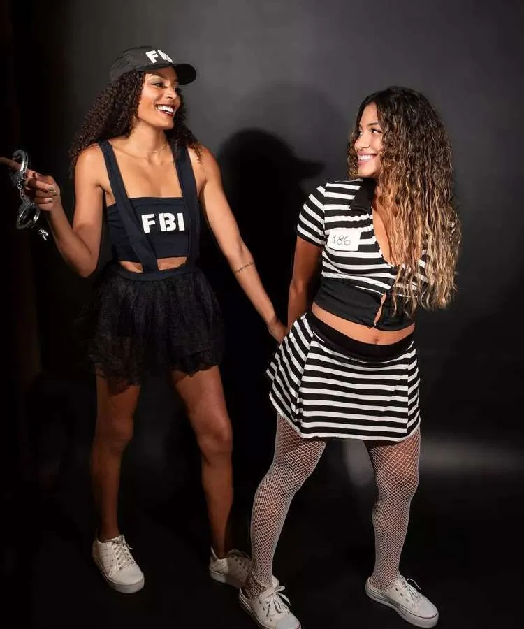 Fantasia de casal para o Halloween: FBI e presidiária. Uma mulher de pele negra usa top preto escrito "FBI" e saia preta, além de boné escrito "FBI" e algemas. A outra mulher usa cropped e saia listrados de branco e preto