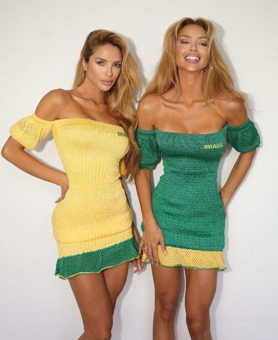 Imagem com fundo de parede branca. No centro, 2 mulheres usando roupa copa do mundo: vestido curto verde e amarelo.