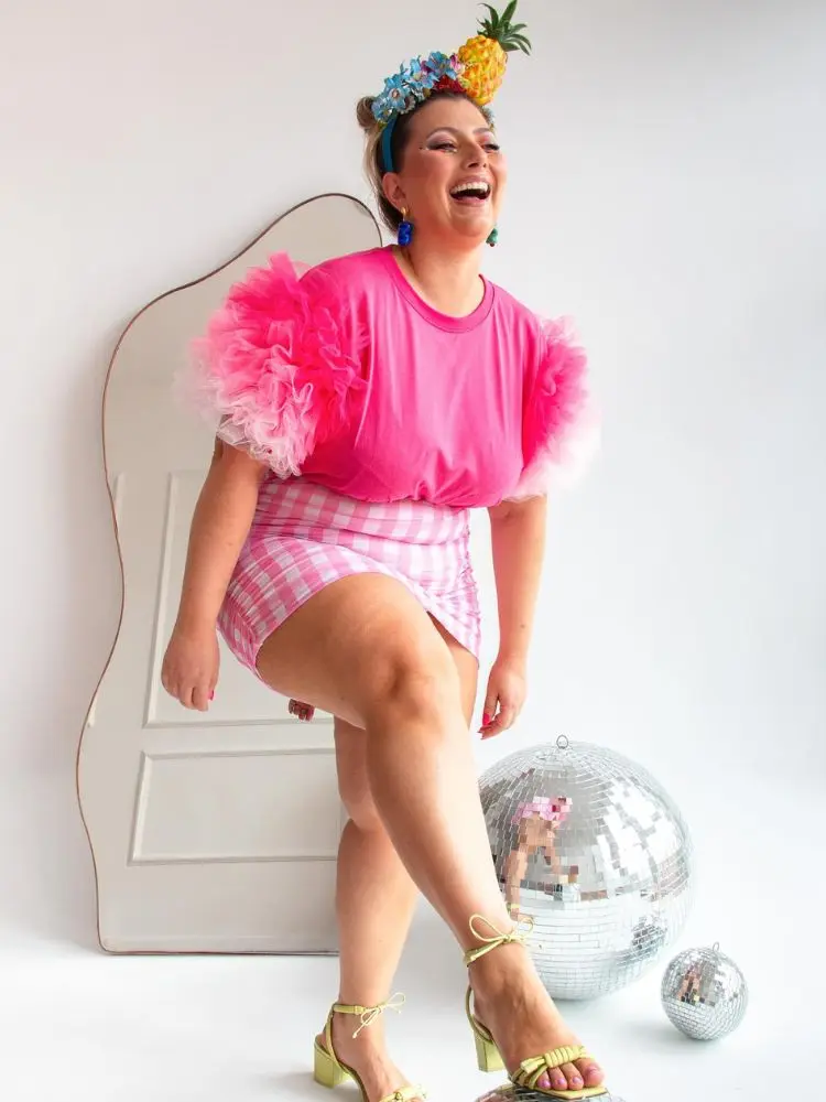 Mulher de pele clara usando blusa rosa com manga de babado de tule, saia xadrez rosa e tiara de frutas 
