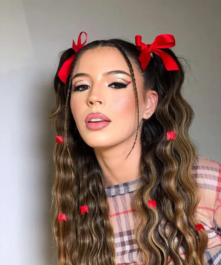 Uma jovem com maquiagem para Festa Junina, apresentando um delineado gráfico preto com detalhes em vermelho, lábios em tom nude-rosado e sardas falsas. Seu cabelo castanho-claro está penteado em marias-chiquinhas com laços vermelhos e fitas vermelhas presas nas tranças.