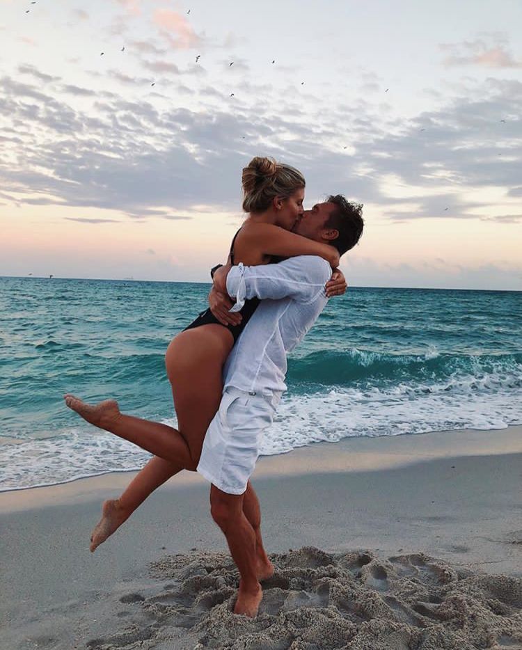 Homem de pele clara usando camisa e bermuda brancas, segurando mulher de pele clara usando maiô, na beira do mar