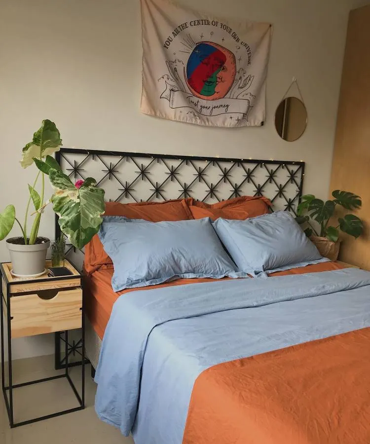 Um quarto com uma cama de lençóis azuis e cobertor laranja. Ao lado, uma alocasia em um vaso cinza está sobre um criado-mudo de madeira. As folhas são grandes e verdes, adicionando um toque natural ao ambiente. Uma tapeçaria colorida decora a parede acima da cama.